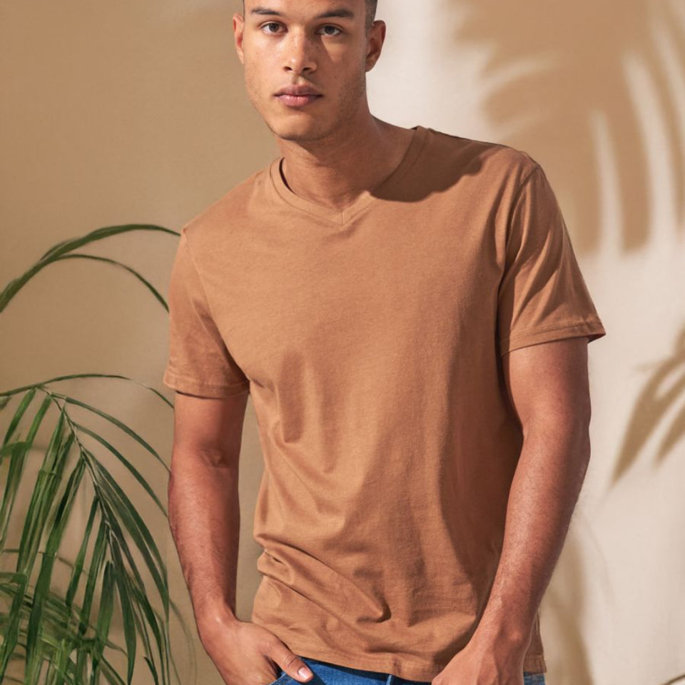 A man wearing a brown v-neck t-shirt and blue jeans, standing in a warm-toned indoor setting with a plant and palm leaf shadows on the wall.