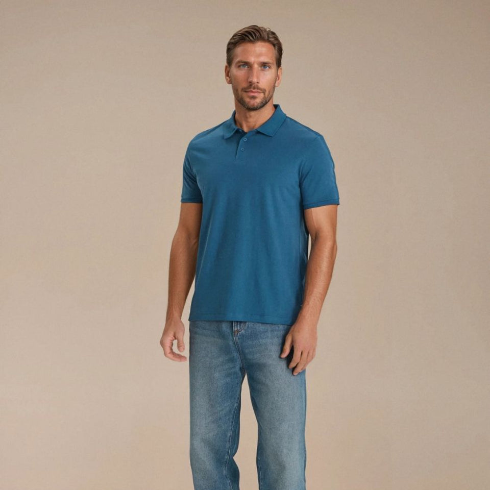 A man modeling a short-sleeve polo shirt in a solid teal blue color, paired with blue jeans and white sneakers, standing against a solid beige studio background.