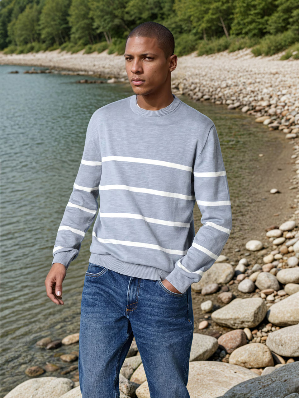 Man wearing a gray sweater with white stripes and blue jeans standing by a body of water.