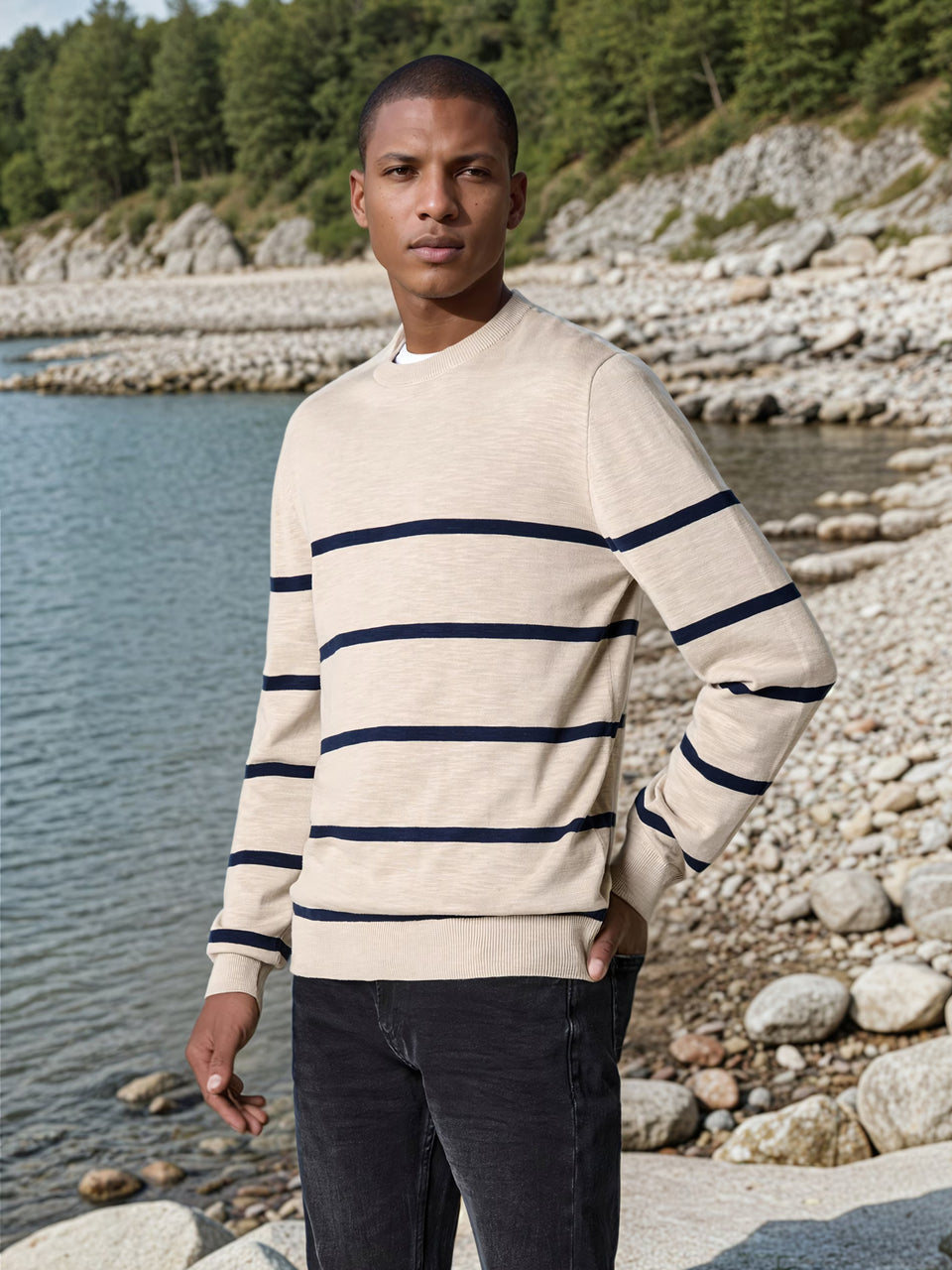 Man wearing a striped sweater standing by a body of water with rocks and trees in the background
