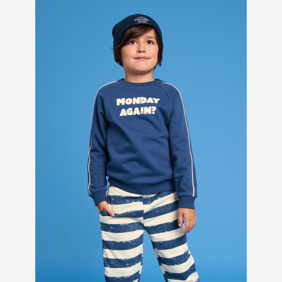 A young boy wearing a navy blue sweatshirt with the text 'MONDAY AGAIN?' in white, matching navy beanie, and navy and white striped pants, standing against a solid blue background.