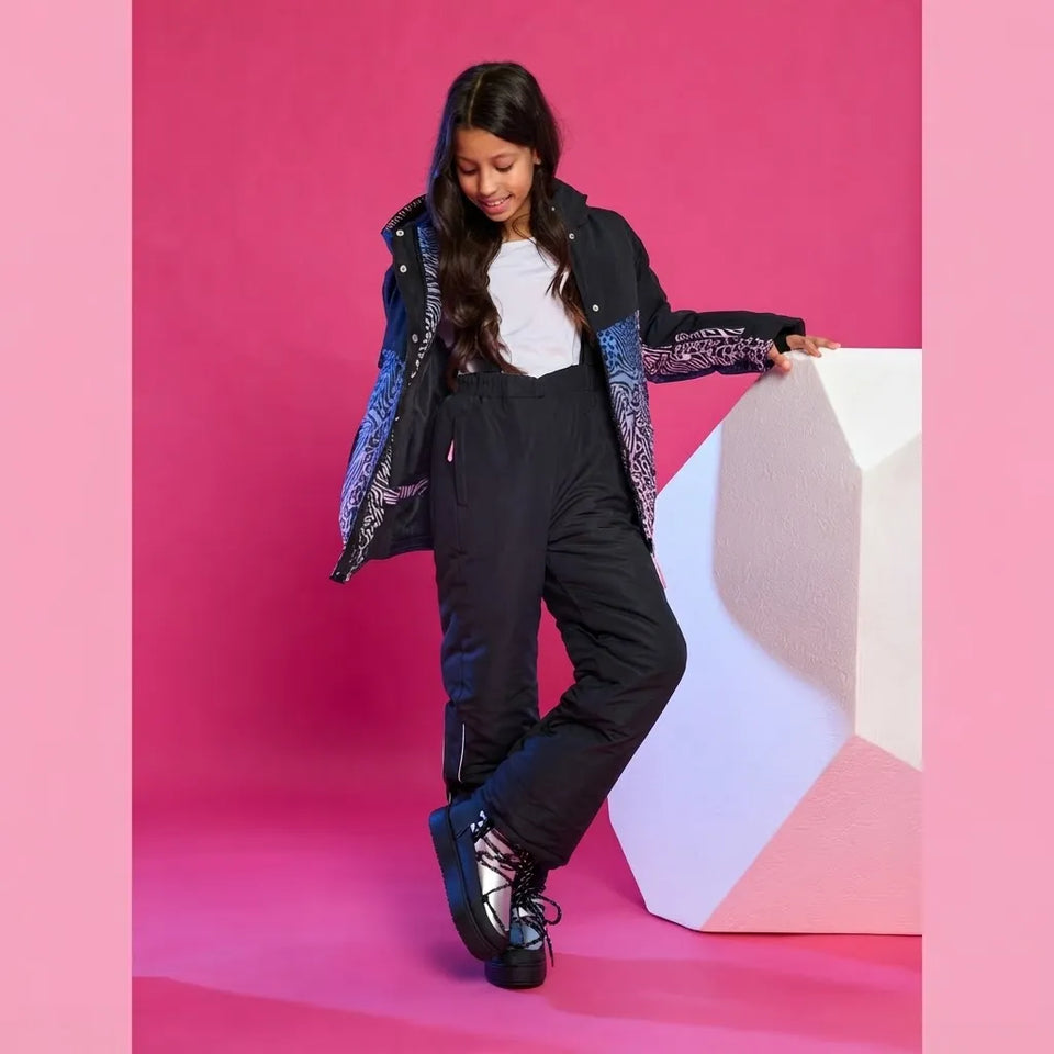 A young girl models a black ski jacket and matching black ski pants with patterned blue and purple accents, posing next to a white geometric block against a vibrant pink background.