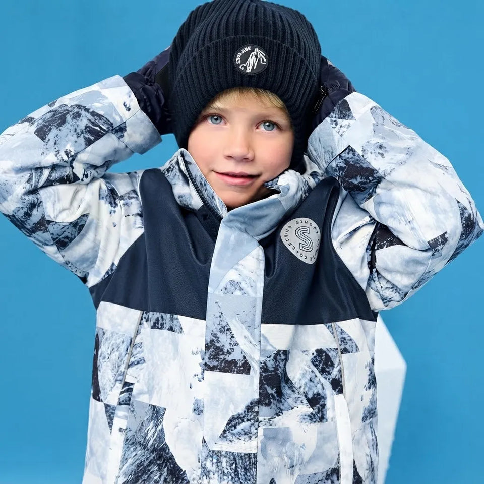 A child wearing a black knit beanie with a white logo and a black-and-white patterned ski jacket, posing against a solid blue background.