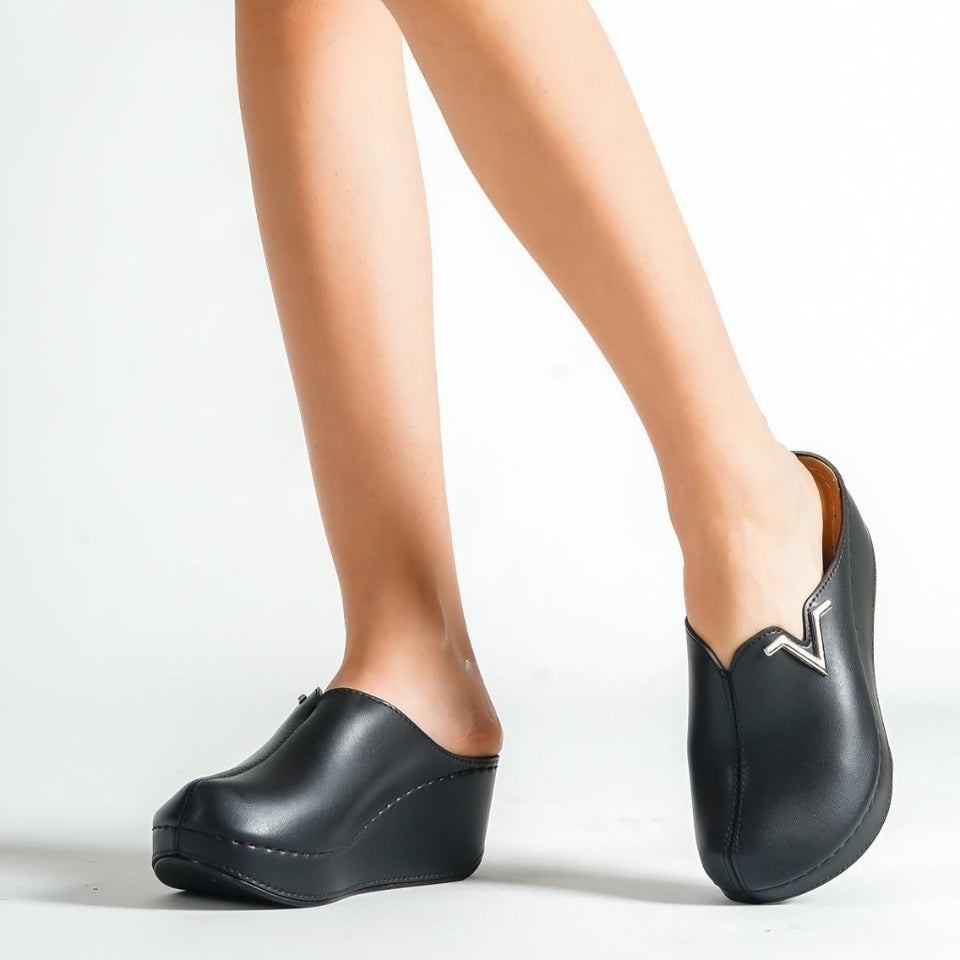 A person wearing black slip-on platform clogs with a small metallic logo on the side, shown from the knees down against a clean white studio background.