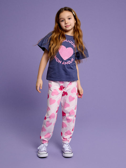 Young girl wearing a navy t-shirt with a pink heart design and white pants with pink heart patterns on a purple background