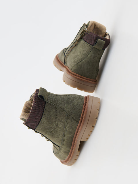 Pair of green suede boots with brown soles and straps on a white background
