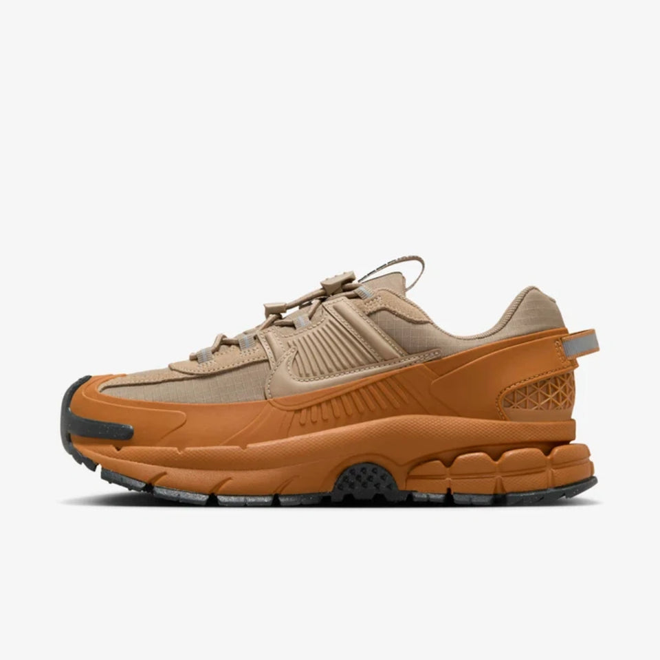 A Nike sneaker in a warm tan and orange colorway, featuring a chunky midsole, textured synthetic overlays, and a black outsole, shown from a side profile on a light gray background.
