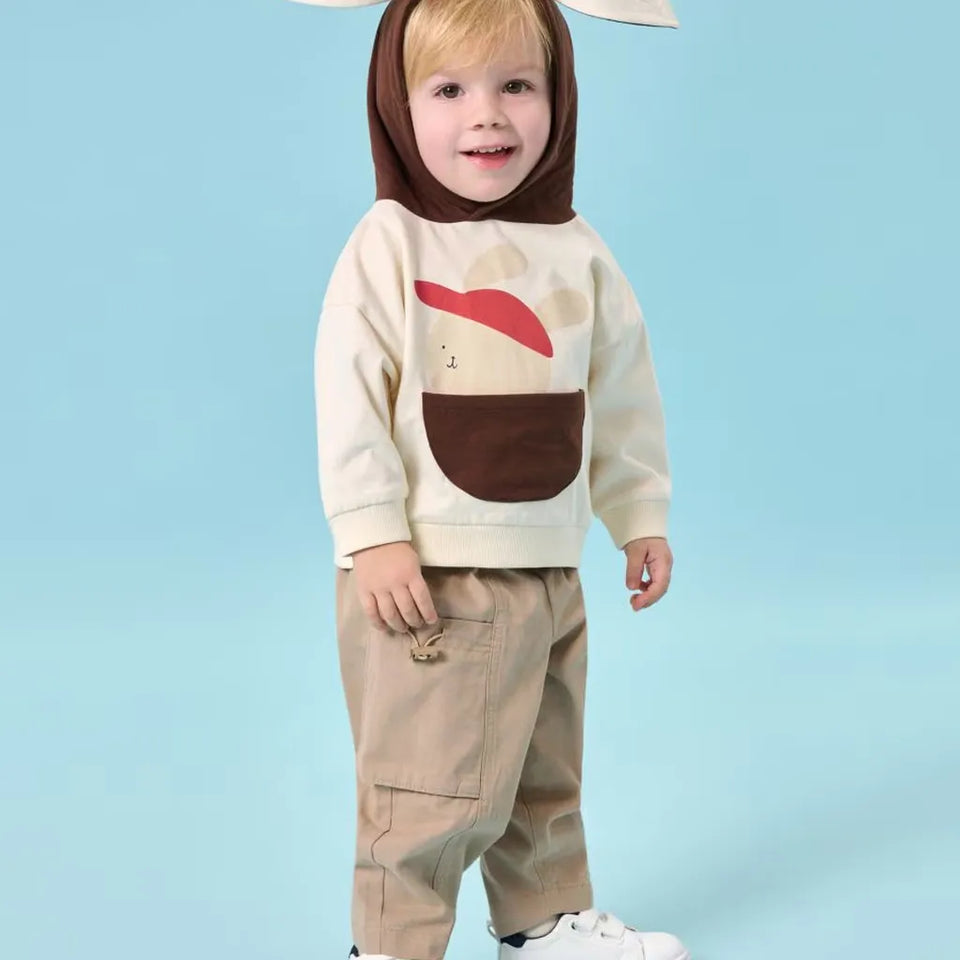 A toddler wearing a beige and brown hooded sweatshirt with a red accent graphic, paired with beige pants, standing against a light blue studio background.