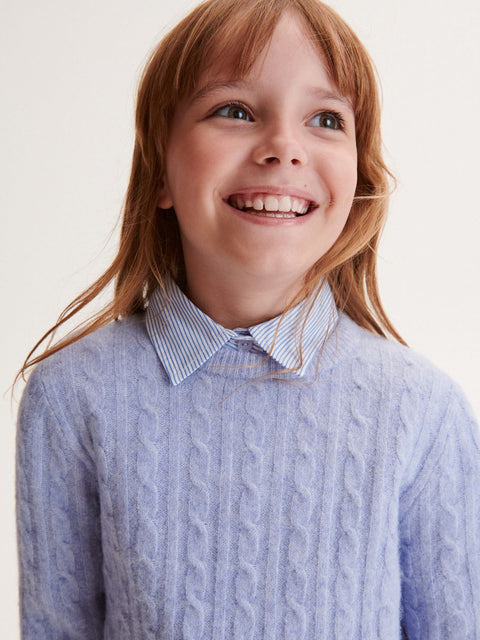 Young girl wearing a light blue cable knit sweater over a collared shirt on a white background