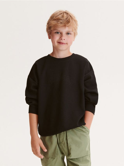 Young boy wearing a black sweater and green pants on a plain background