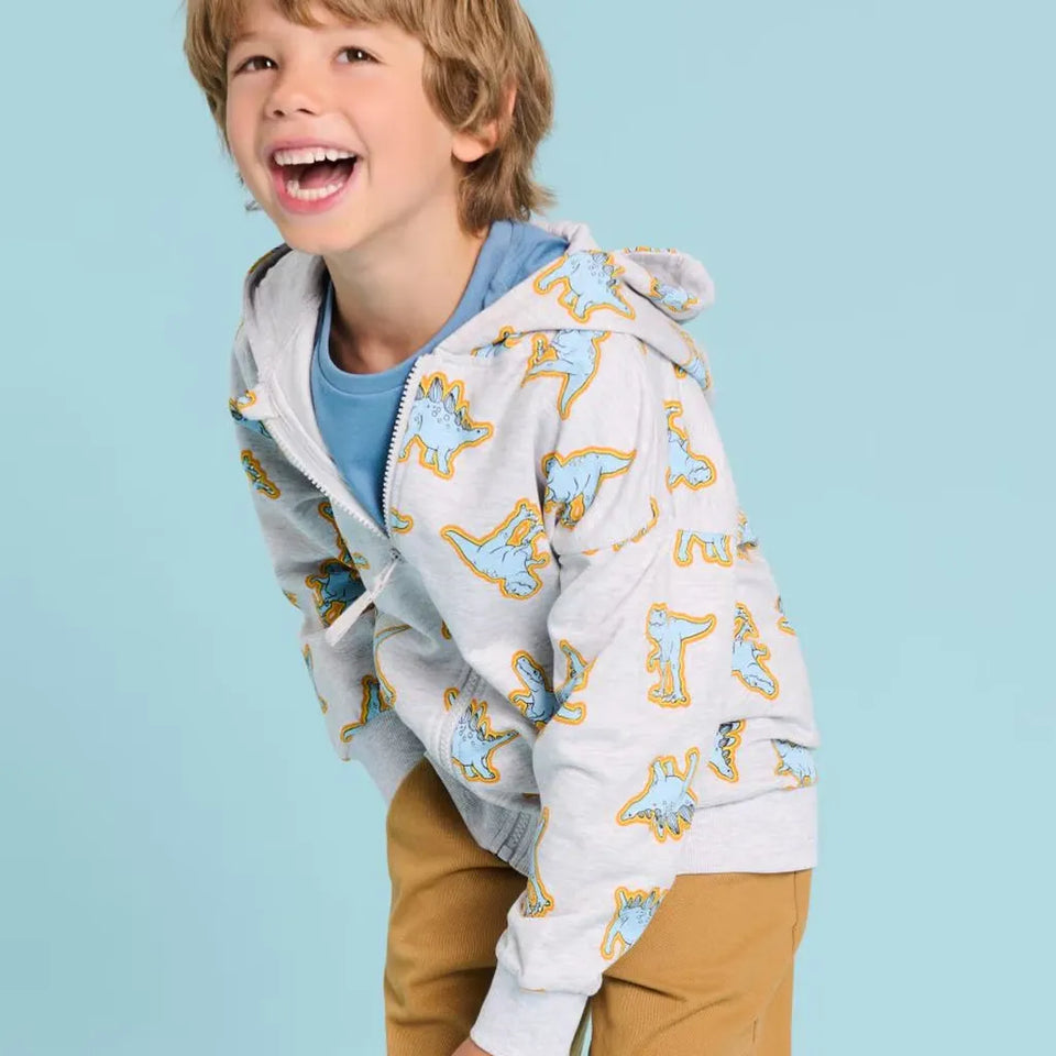 A young boy smiling and looking up, wearing a light gray zip-up hoodie with a blue and yellow dinosaur print, a blue t-shirt, and tan pants, set against a solid light blue background.