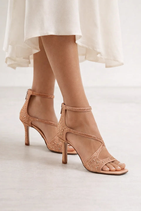 Rose gold high-heeled sandals with intricate detailing worn with a white dress.