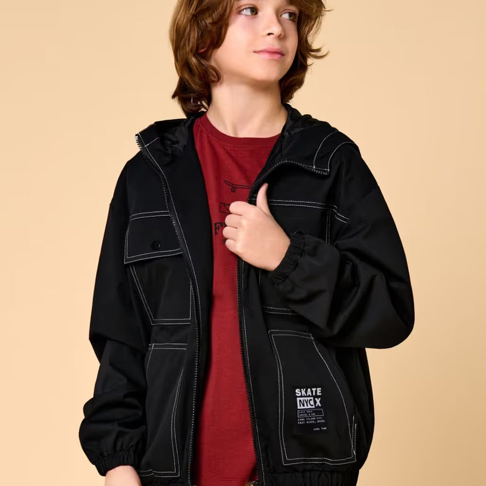 A child wearing a black hooded jacket with white contrast stitching over a red t-shirt, standing against a solid beige background. The jacket features a front zipper, a buttoned chest pocket, and a lower patch pocket with a 'SKATE KICK' brand label.