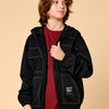 A child wearing a black hooded jacket with white contrast stitching over a red t-shirt, standing against a solid beige background. The jacket features a front zipper, a buttoned chest pocket, and a lower patch pocket with a 'SKATE KICK' brand label.