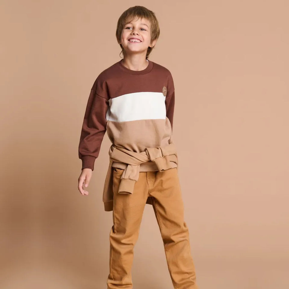 A young boy smiling and posing in a color-blocked sweatshirt with a matching belt and tan pants, set against a solid beige studio background.