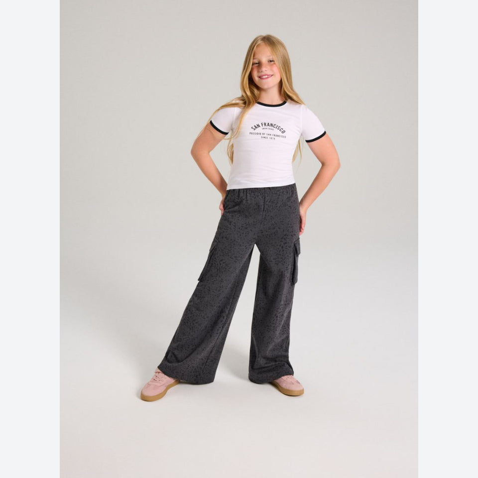 A young girl modeling a white ringer t-shirt with black trim and 'San Francisco' printed on the chest, paired with dark gray wide-leg cargo pants and pink slip-on shoes, against a light gray studio background.