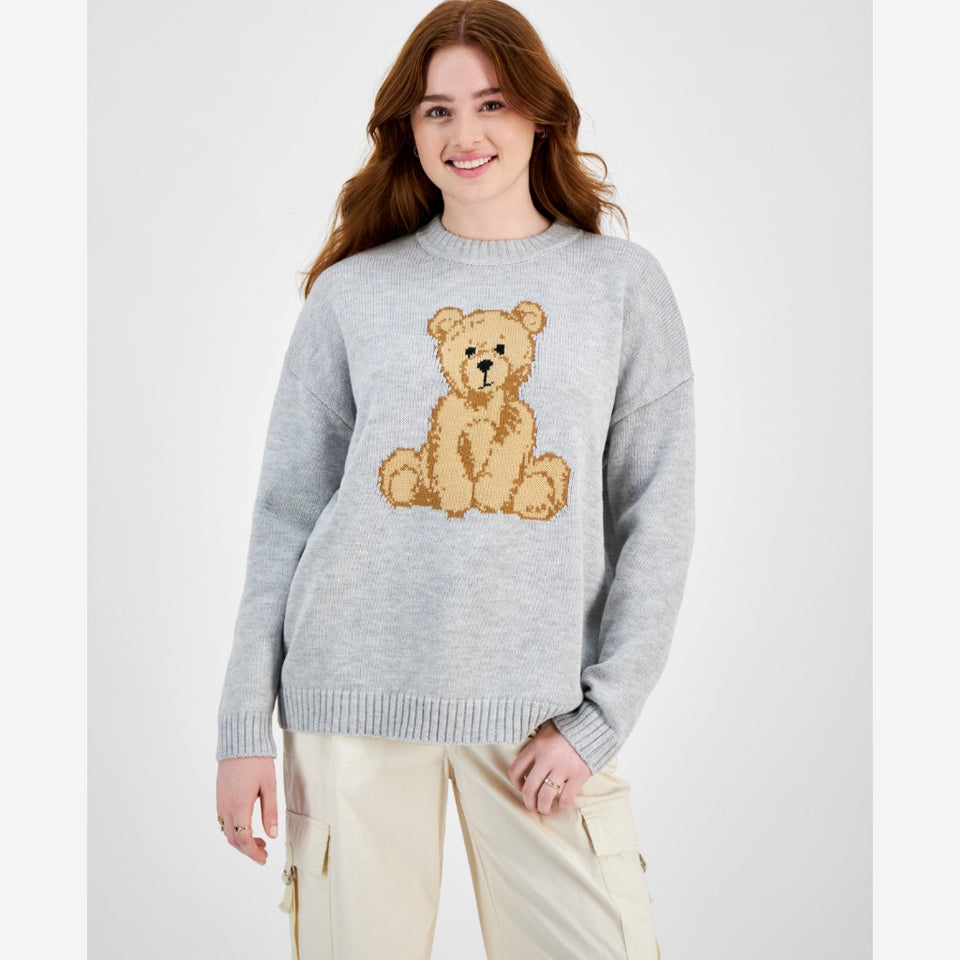 A woman wearing a light gray crewneck sweater with a knitted teddy bear graphic on the chest, paired with beige cargo pants, against a plain light gray background.