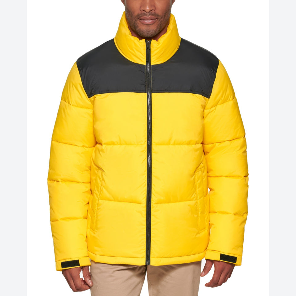A man wearing a bright yellow puffer jacket with a contrasting black yoke across the shoulders and collar, paired with beige pants, shot against a plain white background.