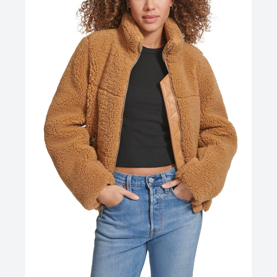 A woman wearing a cropped tan sherpa fleece jacket with a smooth faux-leather front panel, styled over a black crop top and blue jeans against a white studio background.