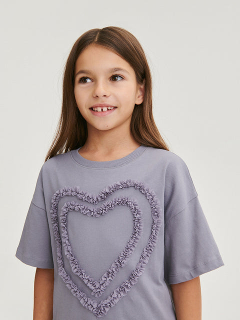 Young girl wearing a gray t-shirt with a heart design on a plain background