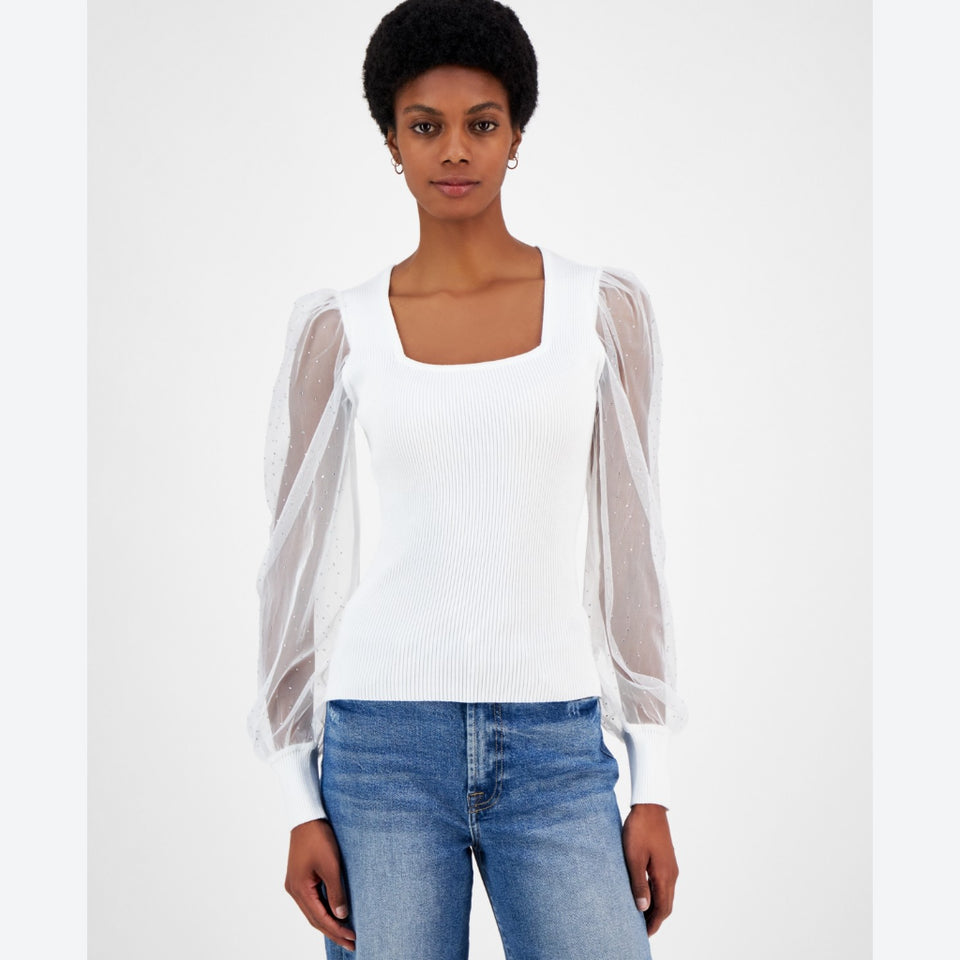 A woman modeling a white ribbed knit top with sheer puff sleeves, paired with blue jeans, against a plain white studio background.