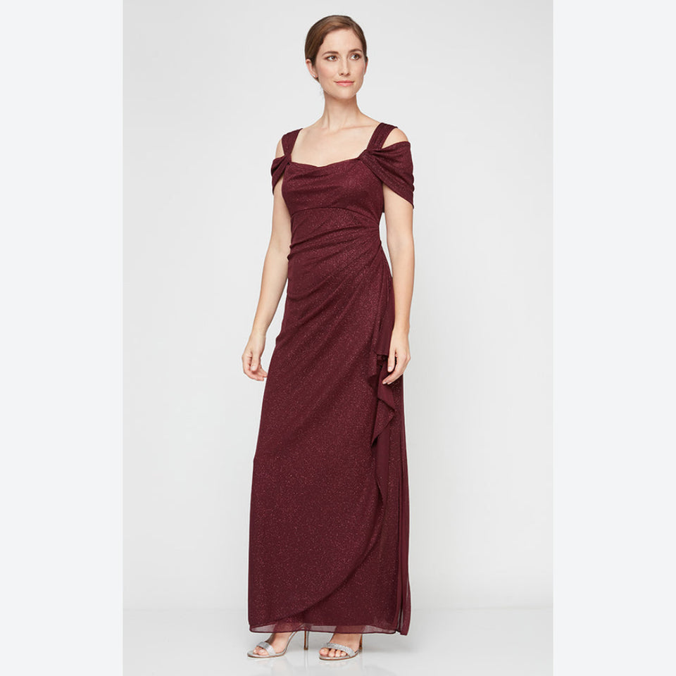 A woman modeling a floor-length burgundy off-the-shoulder gown with a draped, textured fabric and side tie detail, photographed against a light gray studio background.
