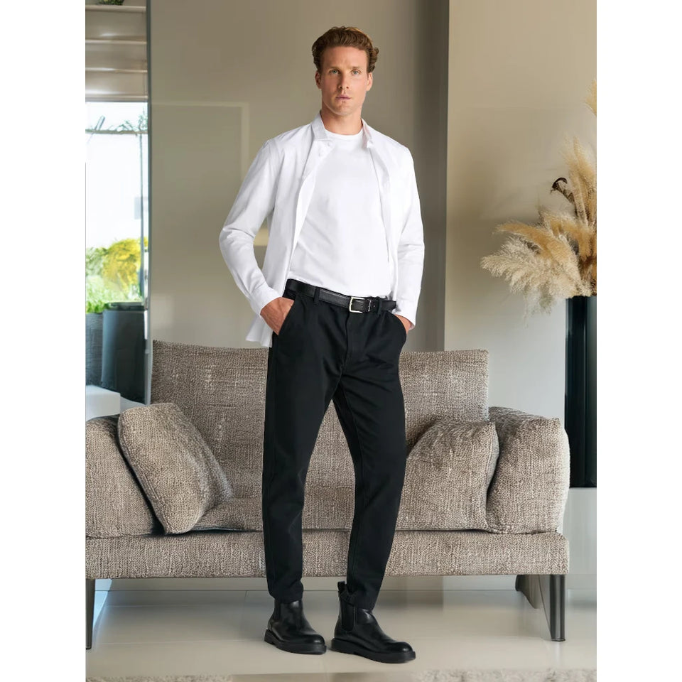 A man stands in a modern living room wearing a white long-sleeve button-down shirt, black dress pants, a black belt, and black boots, posed in front of a textured beige sofa.