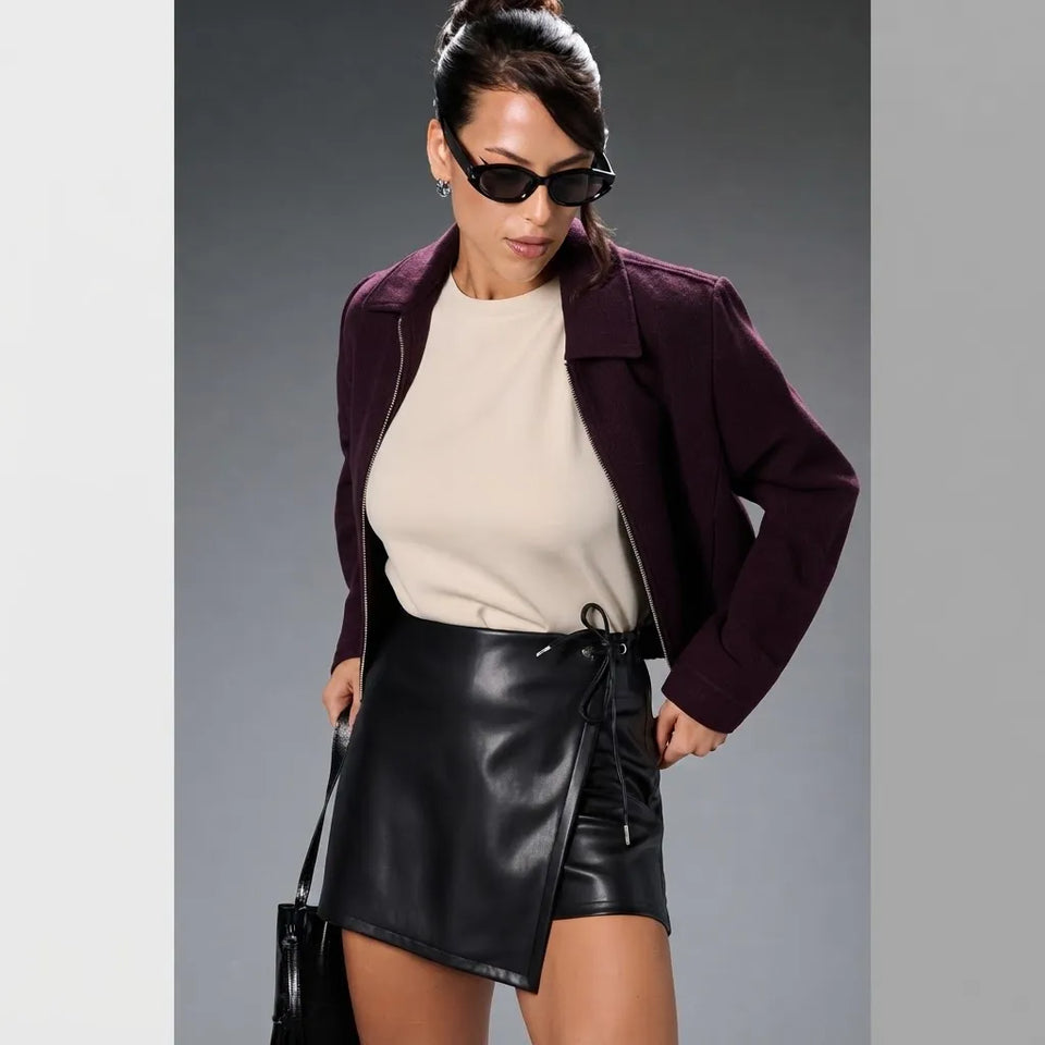 A woman modeling a dark plum corduroy jacket, cream knit top, and black faux-leather wrap mini skirt against a gray studio background.