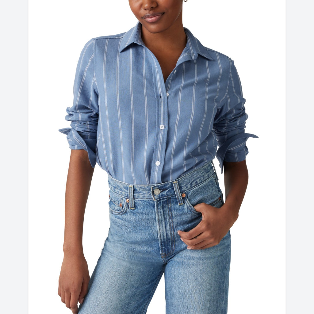 A woman wearing a light blue button-up shirt with thin white vertical stripes and rolled-up sleeves, paired with light-wash blue jeans, shown against a plain white background.