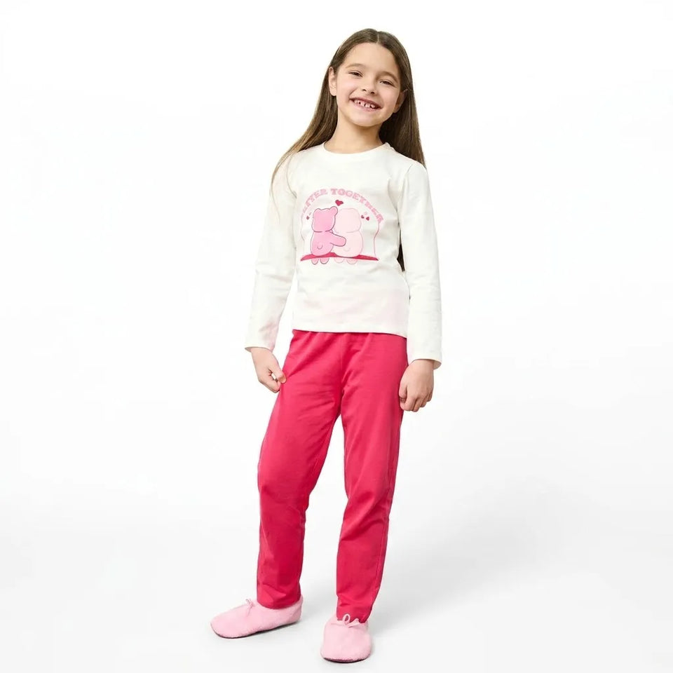 A young girl smiling and wearing a two-piece pajama set consisting of a white long-sleeve top with a pink cartoon bear graphic and bright pink pants, paired with pink slipper shoes, against a plain white background.