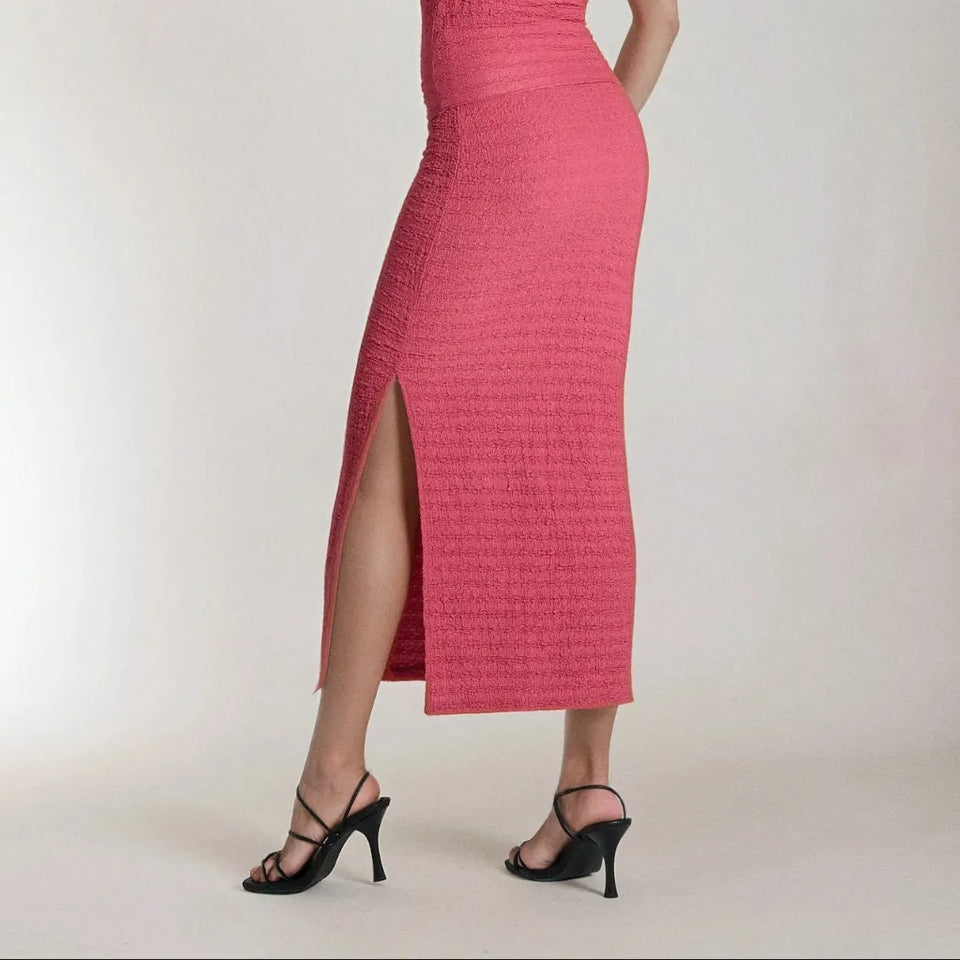 A model is shown from the waist down wearing a textured pink midi skirt with a high side slit and black strappy high-heeled sandals, set against a plain light gray studio background.