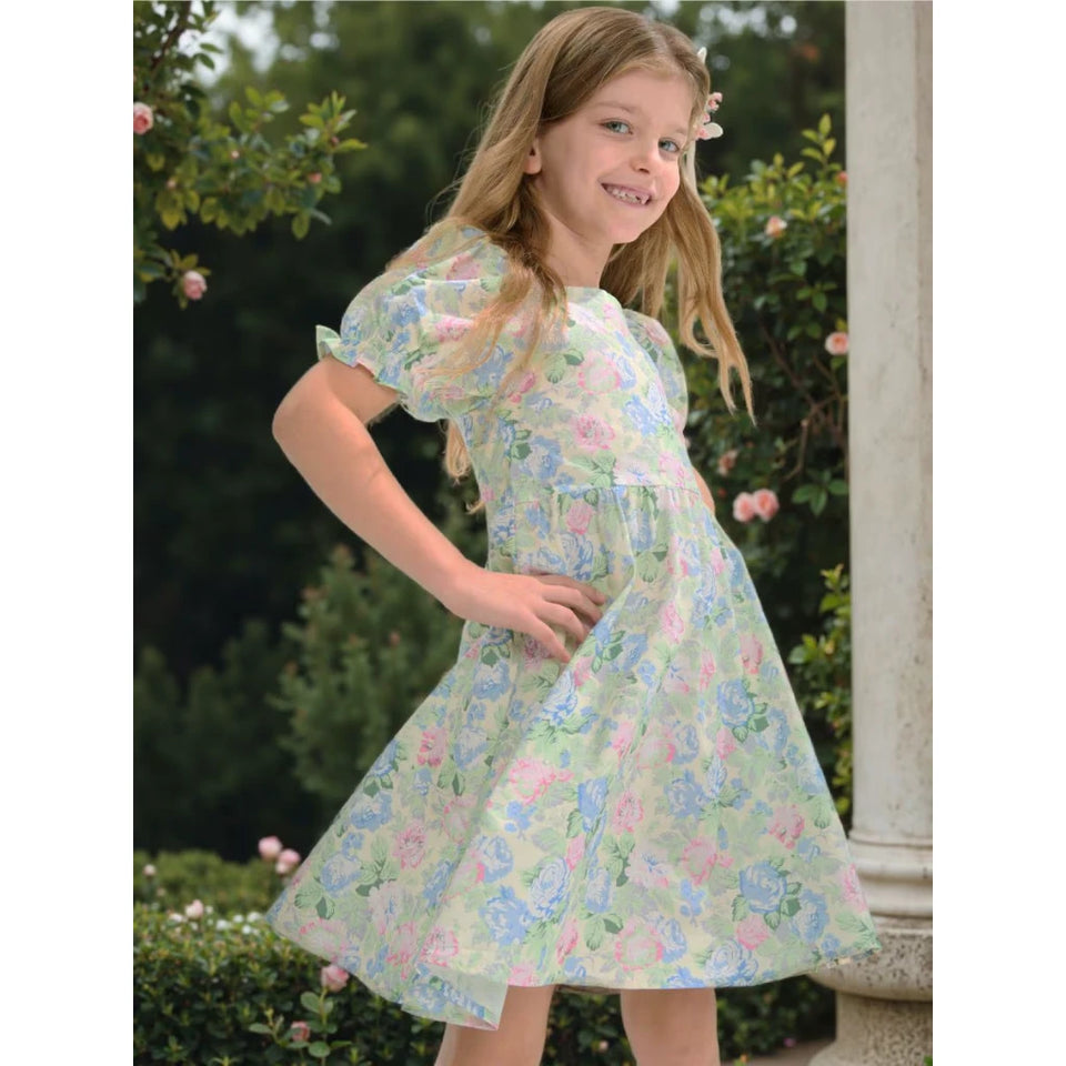A young girl with blonde hair wearing a light green, pink, and blue floral print dress, posing outdoors in front of green bushes and a white pillar.
