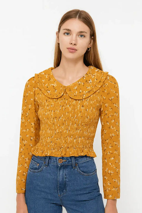 Woman wearing a mustard yellow blouse with a ruffled collar and blue jeans on a white background
