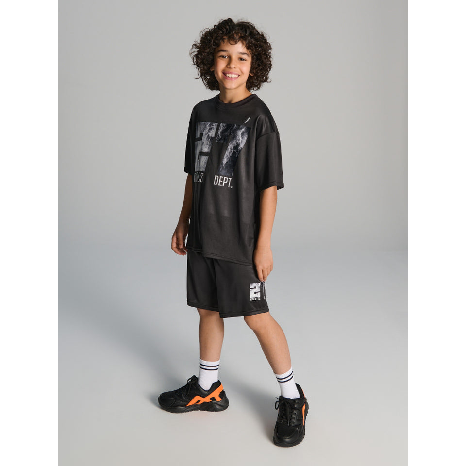 A child model wearing a black graphic t-shirt, matching black athletic shorts, white socks, and black sneakers with orange accents, standing against a neutral gray studio background.
