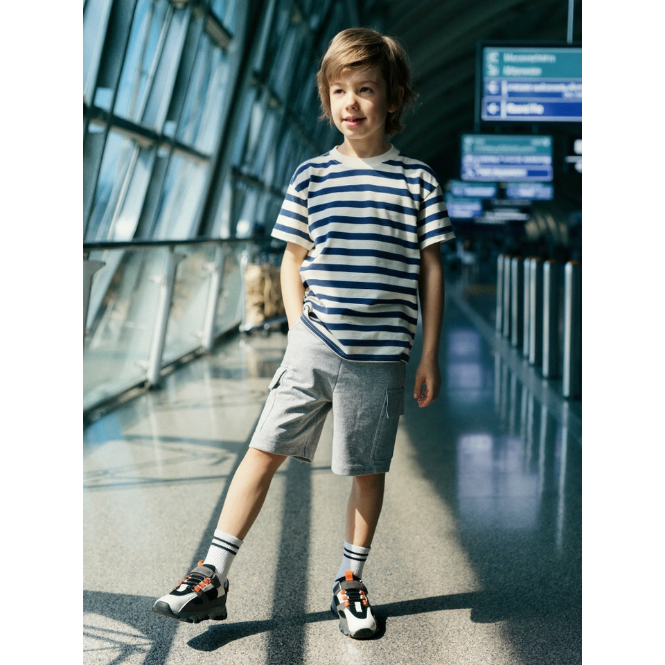 A young boy stands in a brightly lit airport terminal wearing a navy and white horizontally striped short-sleeve t-shirt, light gray shorts, white socks, and black and white sneakers.