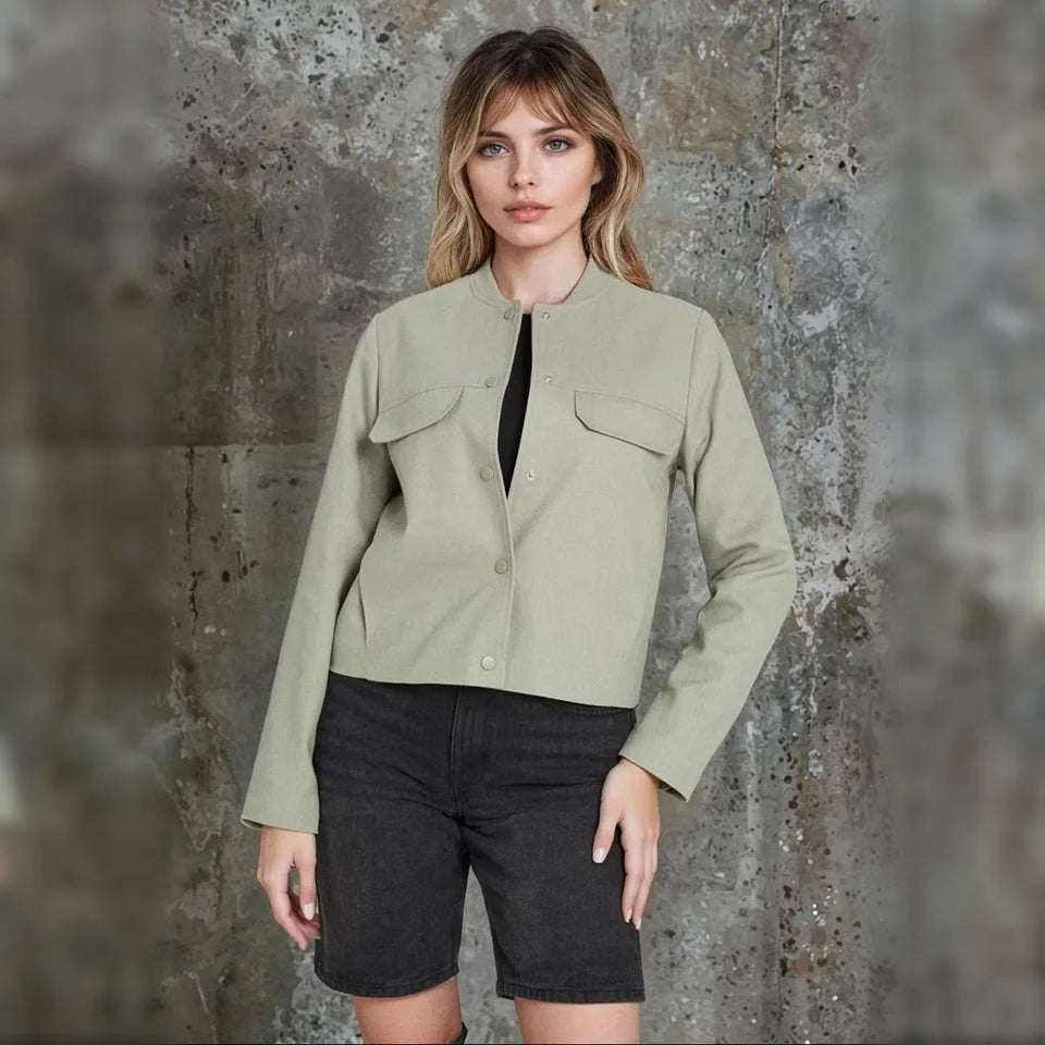 A woman stands against a textured concrete wall wearing a light sage green cropped utility jacket with flap chest pockets over a black top, paired with dark gray denim shorts.