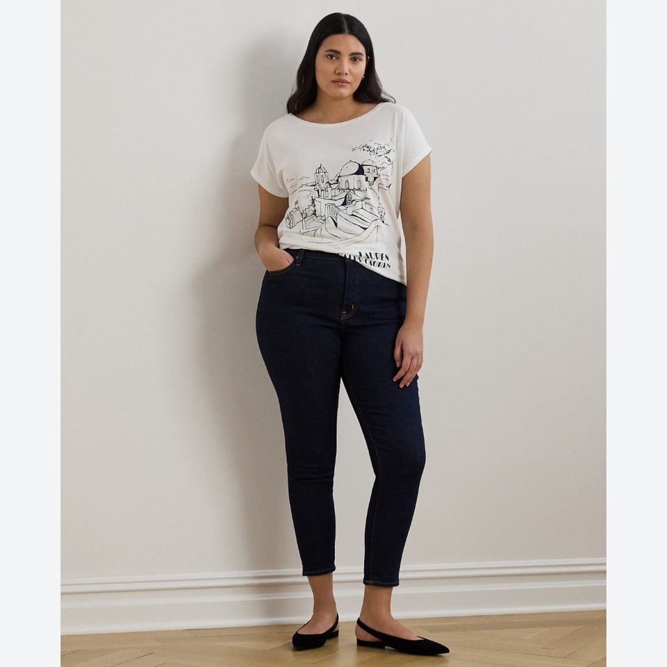A woman stands against a plain off-white wall wearing a white graphic t-shirt, dark navy blue skinny jeans, and black flats.