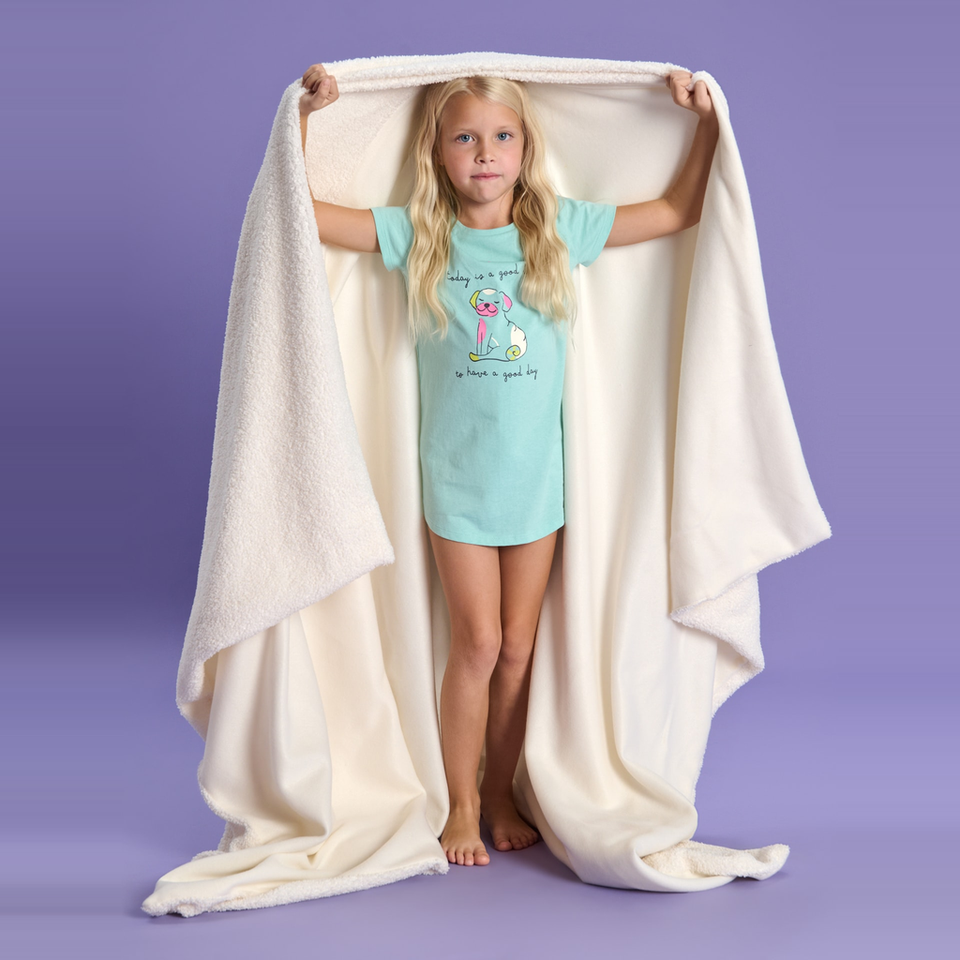 A young girl with blonde hair stands barefoot under a large cream-colored fleece blanket, holding the corners up with her arms. She is wearing a light aqua t-shirt with a cartoon dog graphic and text.