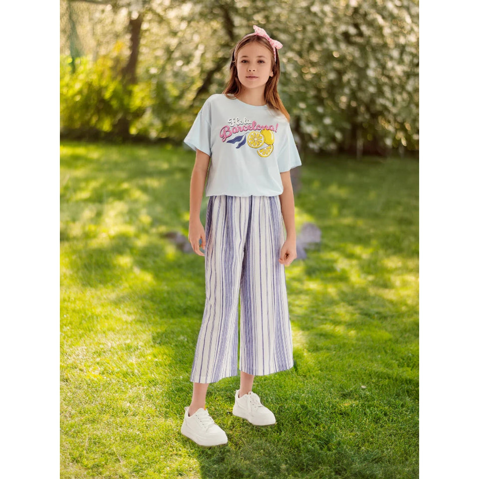 A young girl stands on a grassy lawn wearing a light blue graphic t-shirt, lavender and white striped cropped pants, and white sneakers.