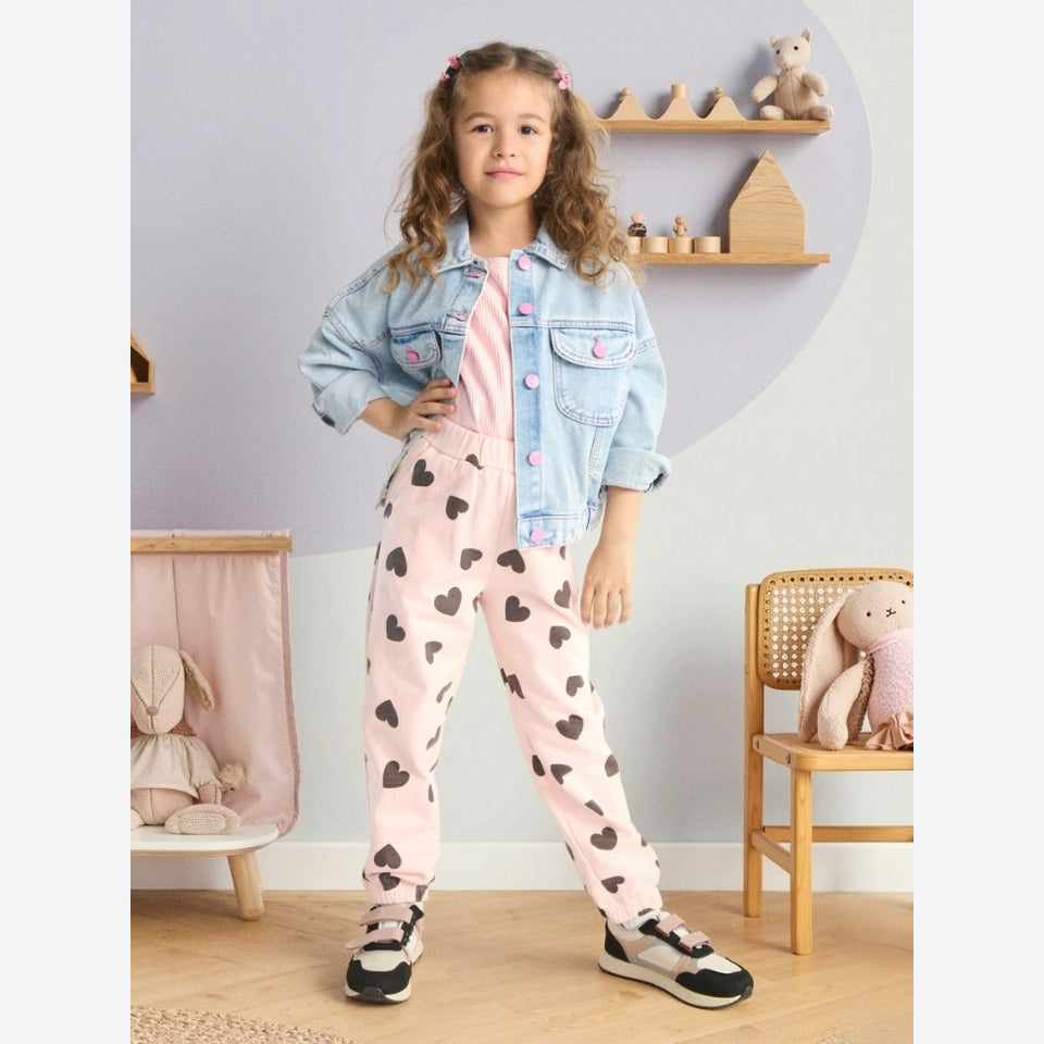 A young girl stands in a child-friendly room wearing a light blue denim jacket over a pink top and pink jogger pants with black heart prints. She is wearing black and white sneakers.