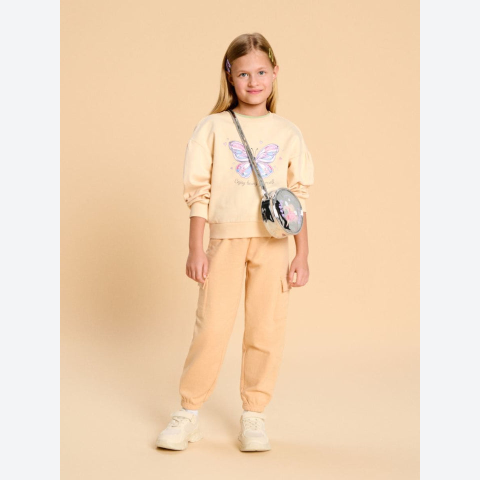 A young girl stands against a solid beige studio background wearing a matching light beige sweatshirt and jogger pants set. The sweatshirt features a colorful butterfly graphic on the front.