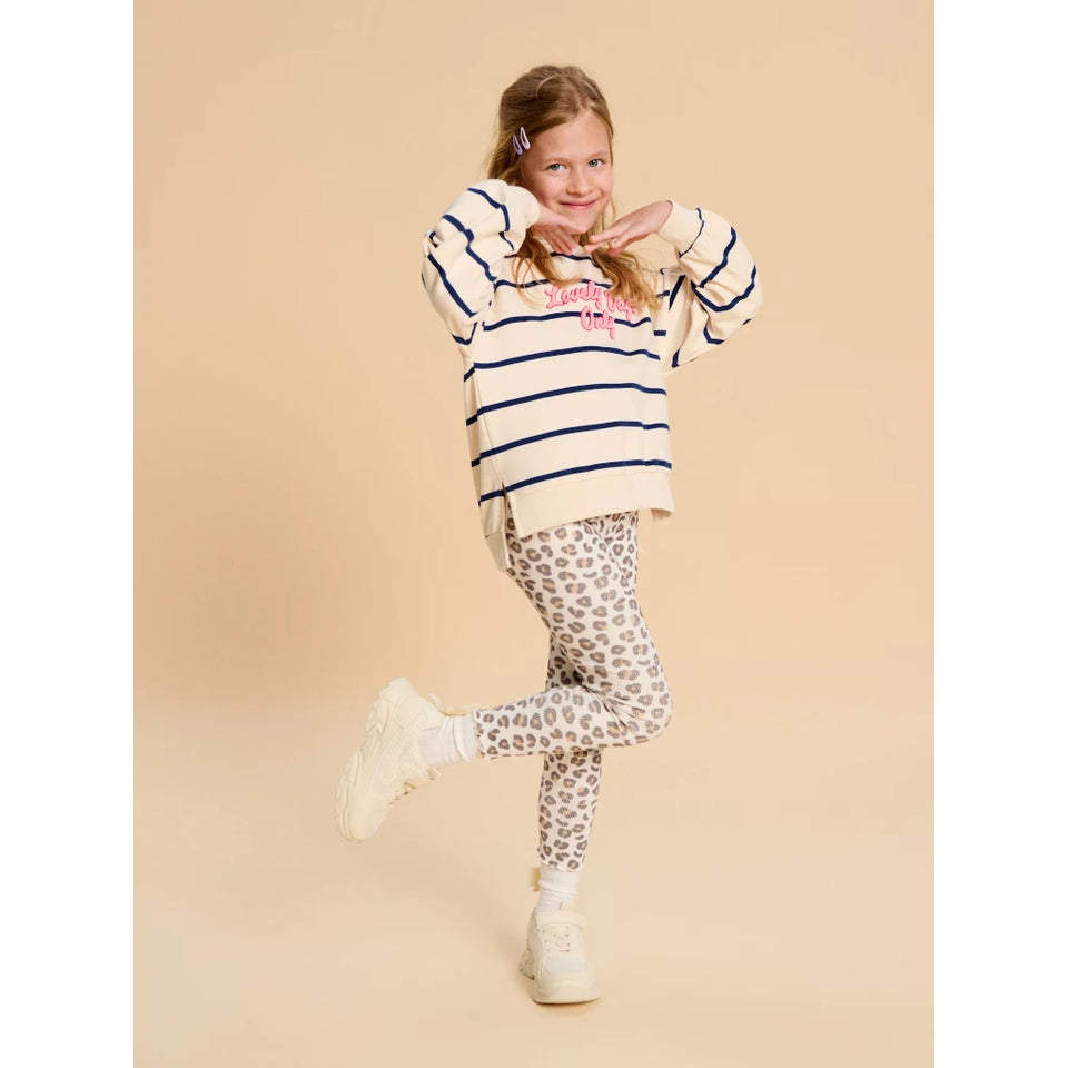 A young girl posing in a cream and navy blue striped knit sweater with pink lettering, paired with leopard print leggings and white sneakers, set against a solid beige background.