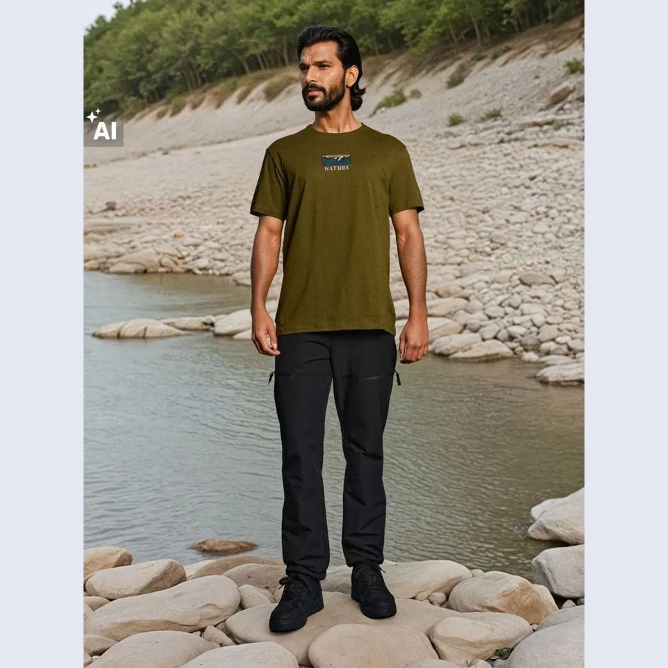 A man stands on rocks by a river wearing an olive green t-shirt with a small blue and white chest graphic, black pants, and black shoes. The background shows a rocky shoreline and green trees.