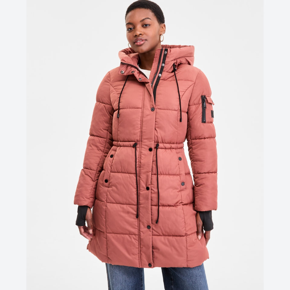 A woman wearing a dusty rose pink hooded puffer coat with black drawstrings and black zipper details, paired with blue jeans, against a plain white studio background.