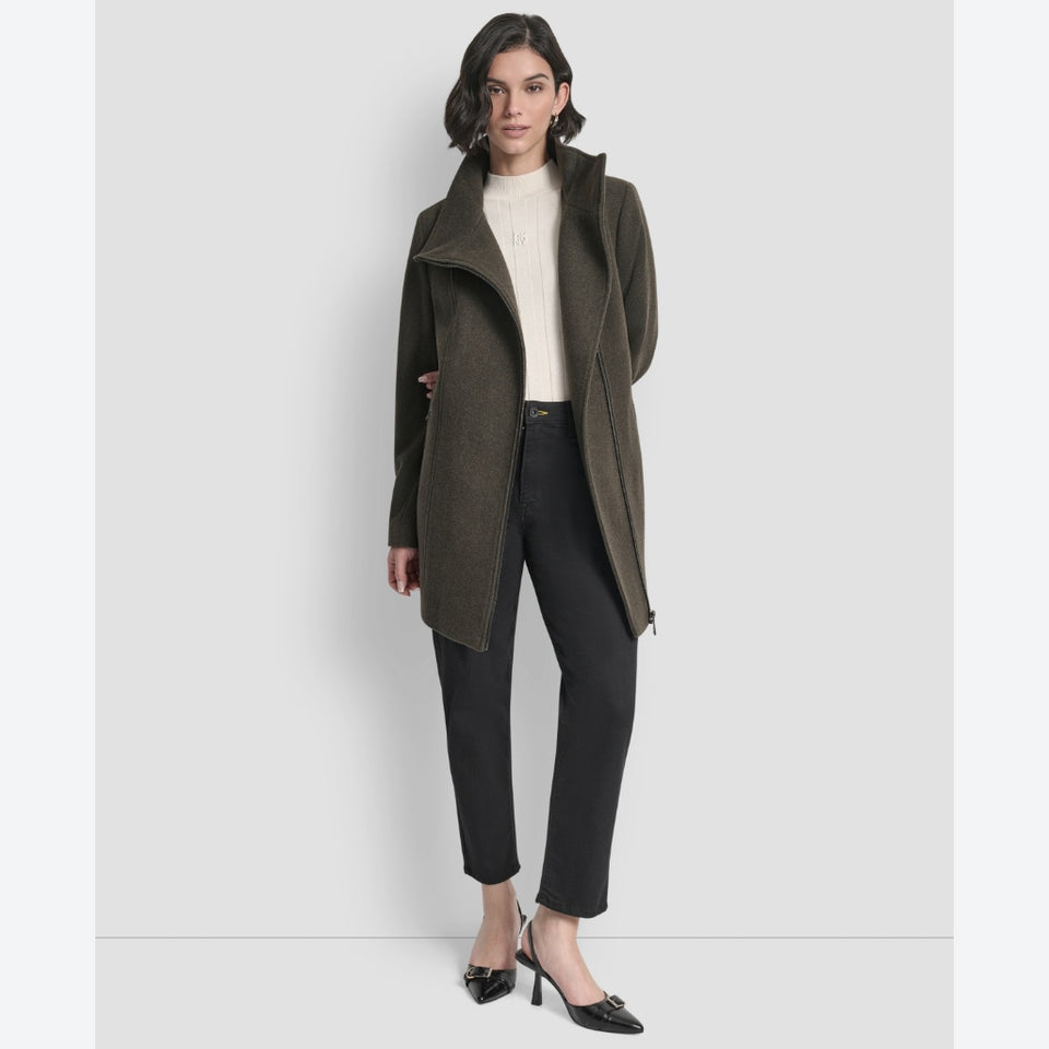 A woman modeling a dark olive green wrap-style coat over a cream ribbed turtleneck, black cropped trousers, and black pointed-toe heels, photographed against a light gray studio background.