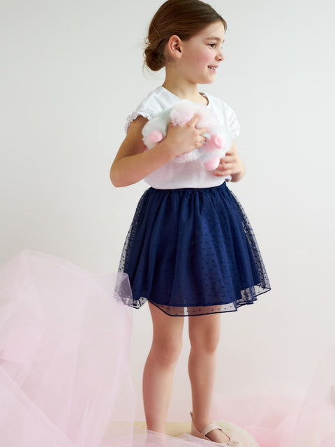 Young girl wearing a white top with pink polka dots and a navy blue skirt on a light background