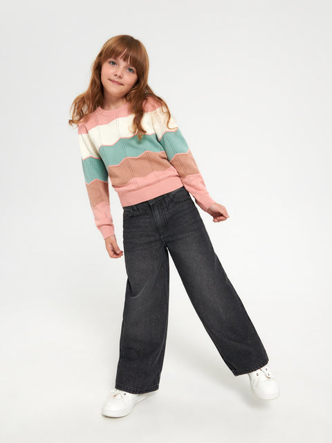 Young girl wearing a colorful striped sweater and dark jeans on a white background
