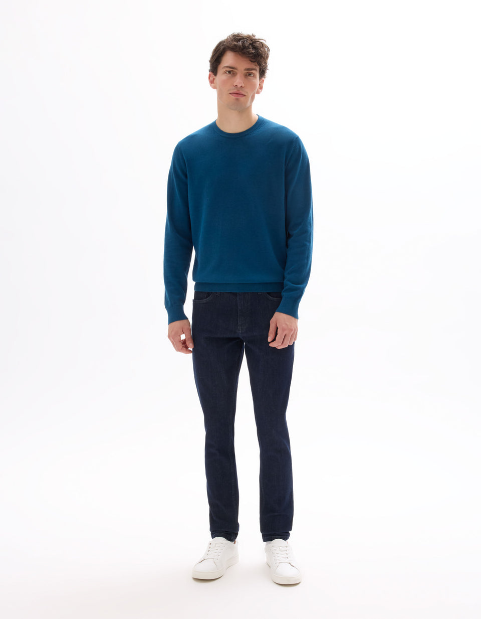Man wearing a blue sweater and dark jeans on a white background