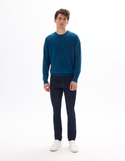 Man wearing a blue sweater and dark jeans on a white background