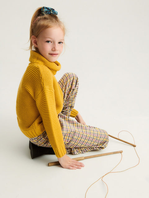 Child wearing a yellow knitted sweater and plaid pants sitting on the floor with knitting materials.